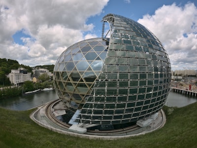 A large, modern spherical building with a partial open structure covered with solar panels sits beside a river. The building's glass and metal design reflects light, and surrounding it are green trees and urban structures. The sky is filled with scattered clouds, adding to the futuristic feel of the setting.