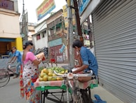 A street scene in an urban area where a fruit vendor is displaying produce on a cart. A woman in a colorful saree is examining the fruits while wearing a mask. The street is lined with shops, one of which has a closed shutter. Another person is riding a bicycle in the background. The environment appears busy but subdued.