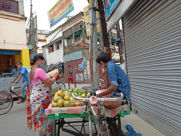 A street scene in an urban area where a fruit vendor is displaying produce on a cart. A woman in a colorful saree is examining the fruits while wearing a mask. The street is lined with shops, one of which has a closed shutter. Another person is riding a bicycle in the background. The environment appears busy but subdued.