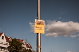 A pole with a yellow sign in German that says 'Deutsche Wohnen & Co enteignen!', 'Hier unterschreiben' against a backdrop of a partly cloudy sky. Buildings with red-roof tiles and green trees are visible in the background.