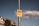 A pole with a yellow sign in German that says 'Deutsche Wohnen & Co enteignen!', 'Hier unterschreiben' against a backdrop of a partly cloudy sky. Buildings with red-roof tiles and green trees are visible in the background.
