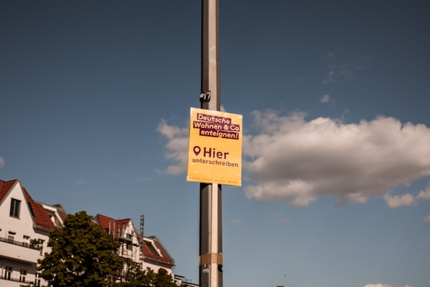 A pole with a yellow sign in German that says 'Deutsche Wohnen & Co enteignen!', 'Hier unterschreiben' against a backdrop of a partly cloudy sky. Buildings with red-roof tiles and green trees are visible in the background.