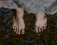A model walking barefoot on grass wearing a lilac linen blouse, surrounded by wildflowers.