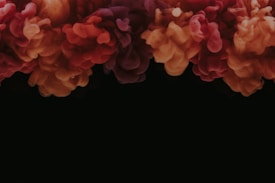 A vibrant cloud of colorful ink or smoke billows against a dark background, creating a striking contrast. The mix of colors includes deep reds, oranges, and purples that blend seamlessly, giving the appearance of a dynamic and flowing substance. The texture appears soft and billowy, resembling swirling clouds.