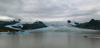 A vast landscape featuring a large glacier extending across rugged mountains under a thick cloud cover. The glacier appears to be slowly flowing into a calm body of water in the foreground. On the left, chunks of ice seem to have broken off from the glacier, floating in the water. A small motorboat is visible on the right, creating gentle ripples as it moves across the water.