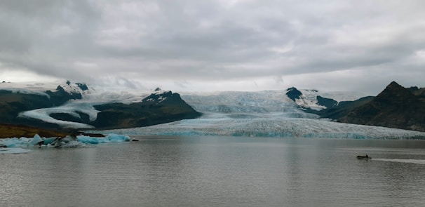 A vast landscape featuring a large glacier extending across rugged mountains under a thick cloud cover. The glacier appears to be slowly flowing into a calm body of water in the foreground. On the left, chunks of ice seem to have broken off from the glacier, floating in the water. A small motorboat is visible on the right, creating gentle ripples as it moves across the water.