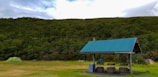 A grassy campsite surrounded by dense, lush green trees under a partly cloudy sky. Two tents are set up on either side, one green and the other orange, with picnic tables nearby. A shelter with a corrugated metal roof stands prominently in the foreground, providing cover for a few stone seats and a bin.
