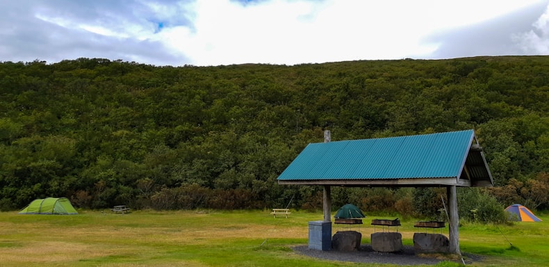 A grassy campsite surrounded by dense, lush green trees under a partly cloudy sky. Two tents are set up on either side, one green and the other orange, with picnic tables nearby. A shelter with a corrugated metal roof stands prominently in the foreground, providing cover for a few stone seats and a bin.