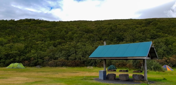 A grassy campsite surrounded by dense, lush green trees under a partly cloudy sky. Two tents are set up on either side, one green and the other orange, with picnic tables nearby. A shelter with a corrugated metal roof stands prominently in the foreground, providing cover for a few stone seats and a bin.