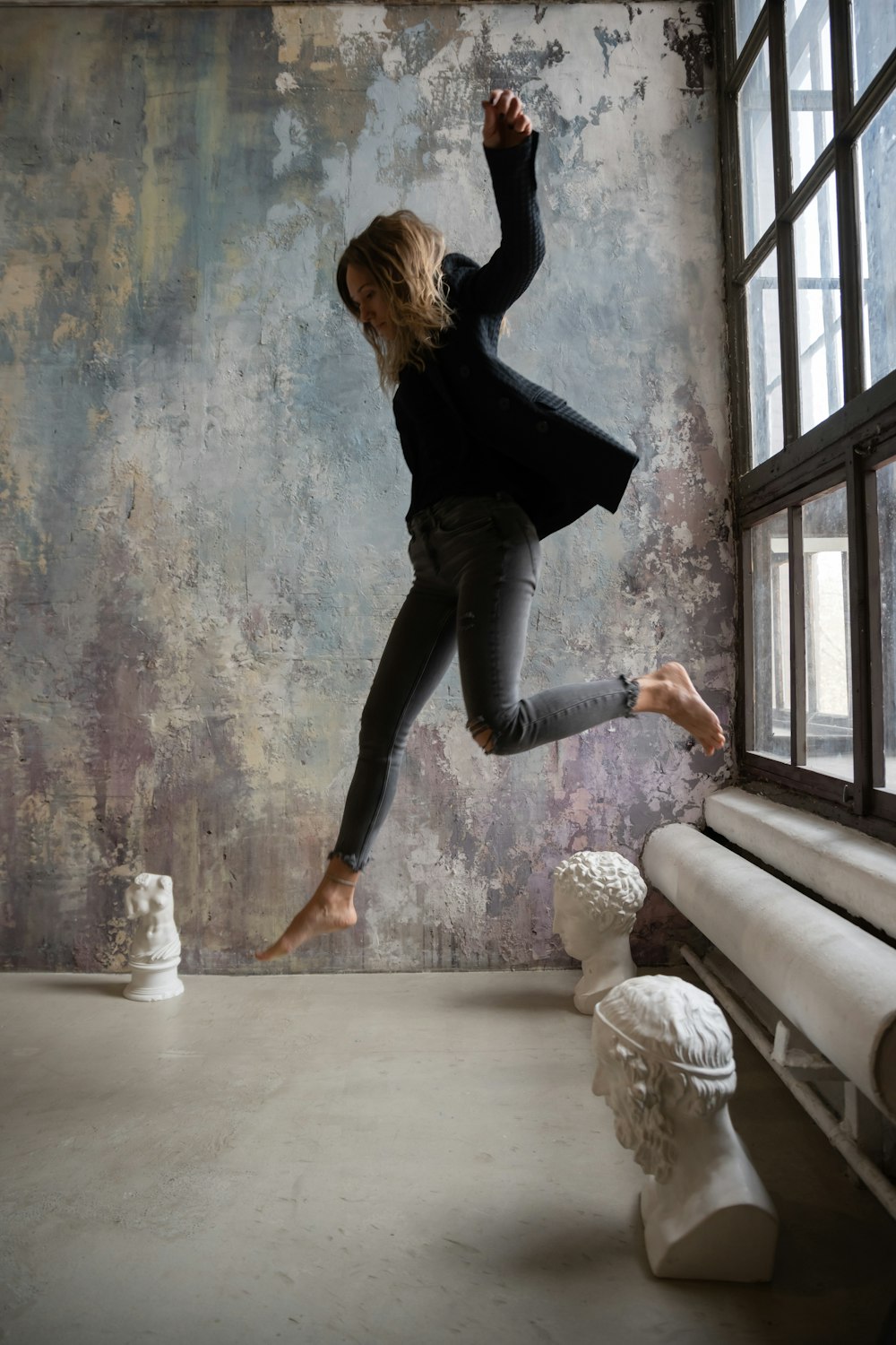 woman in black long sleeve shirt and blue denim jeans jumping on white concrete floor