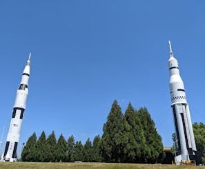 Two large rockets stand upright on a grassy area. The rockets are designed with a white and black color scheme and feature the words 'USA' and 'UNITED STATES' on their sides. They are surrounded by a row of tall green trees, under a clear blue sky.