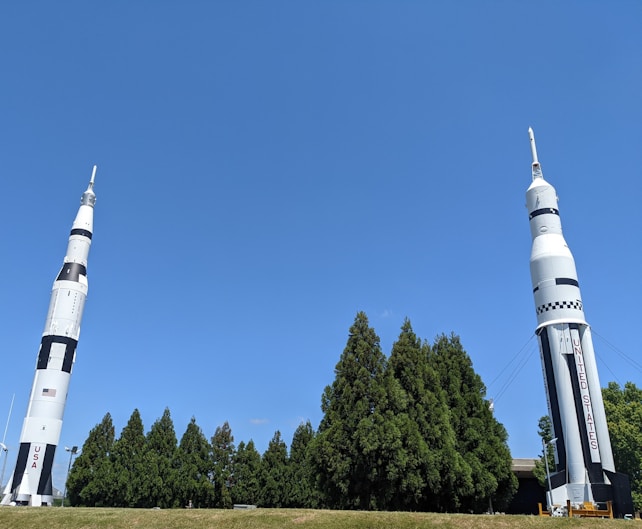 Two large rockets stand upright on a grassy area. The rockets are designed with a white and black color scheme and feature the words 'USA' and 'UNITED STATES' on their sides. They are surrounded by a row of tall green trees, under a clear blue sky.