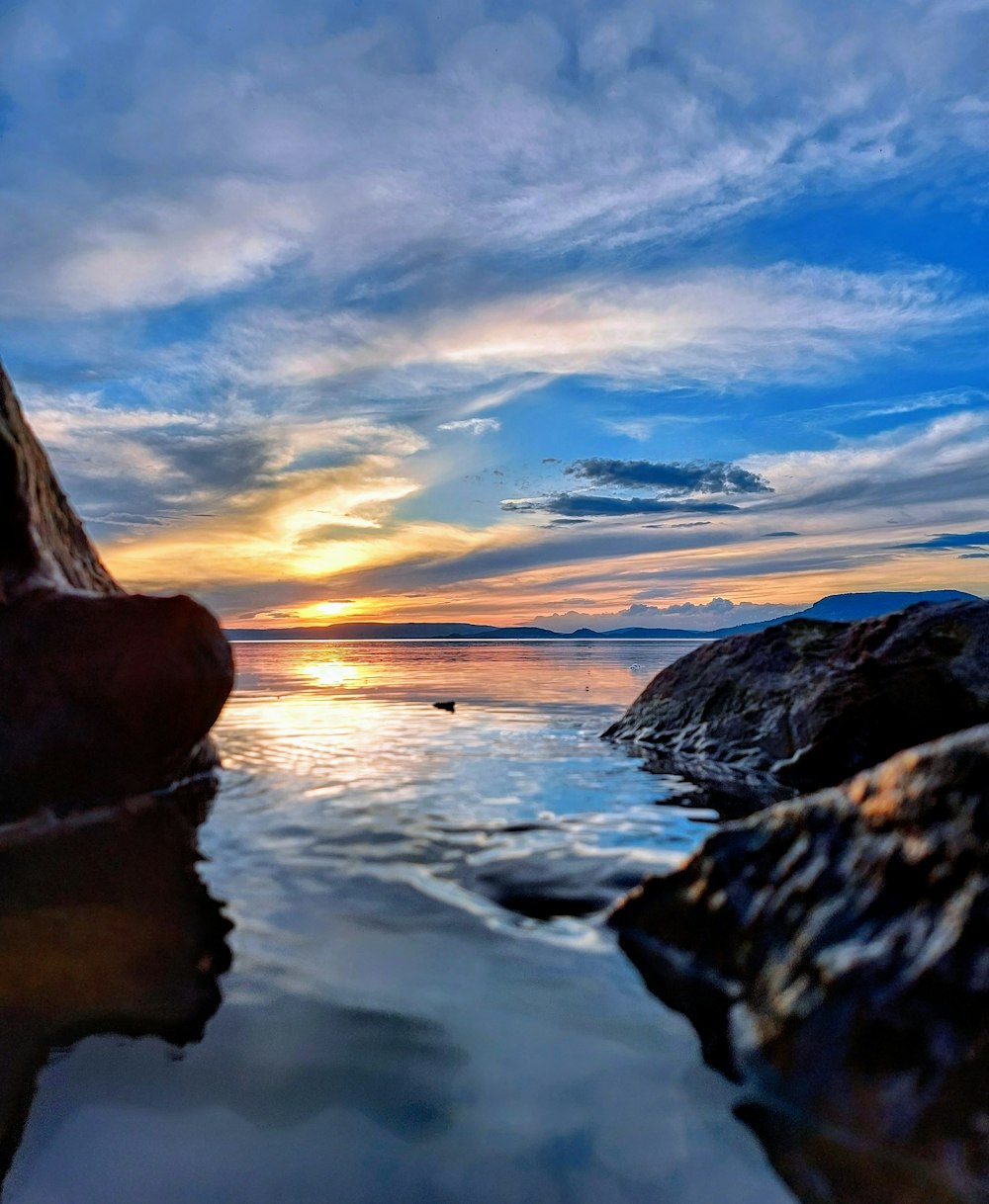 a body of water with rocks in the foreground and a sunset in the background