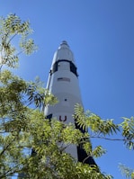 A large rocket stands tall against a clear blue sky, partially obscured by the green leaves of a nearby tree. The rocket is marked with an American flag and features black and white sections.