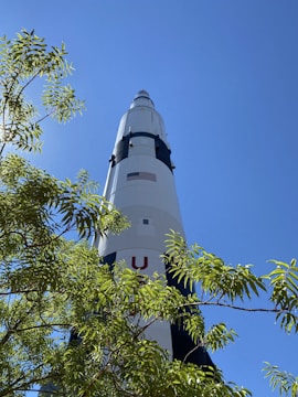 A large rocket stands tall against a clear blue sky, partially obscured by the green leaves of a nearby tree. The rocket is marked with an American flag and features black and white sections.