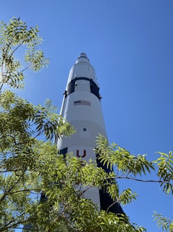 A large rocket stands tall against a clear blue sky, partially obscured by the green leaves of a nearby tree. The rocket is marked with an American flag and features black and white sections.