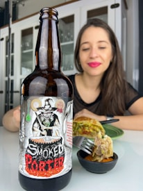 A dark beer bottle with a label featuring a witch stirring a cauldron is prominently displayed on a table. In the background, a woman with bright pink lipstick is smiling softly, and there is a plate with food, possibly a savory dish, being eaten with a fork.