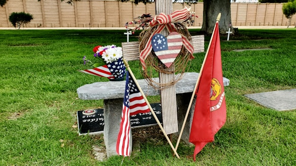 A wooden cross adorned with a heart-shaped decoration featuring the American flag patterns is set against a green grassy backdrop. To the left is an American flag, next to a bouquet of red, white, and blue flowers. A red flag with a military emblem is on the right. Behind the cross is a stone bench engraved with writing, and in the background are additional crosses and a wall.
