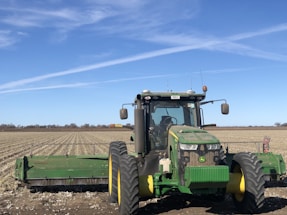 A large green tractor is positioned in the foreground of an expansive, flat farmland under a clear blue sky. The tractor is equipped with wide wheels and farming attachments in the back. The field appears to be recently harvested or prepared for planting, with rows stretching into the distance. The horizon is visible with a few trees far away, and contrails are faintly visible in the sky.