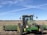 A large green tractor is positioned in the foreground of an expansive, flat farmland under a clear blue sky. The tractor is equipped with wide wheels and farming attachments in the back. The field appears to be recently harvested or prepared for planting, with rows stretching into the distance. The horizon is visible with a few trees far away, and contrails are faintly visible in the sky.