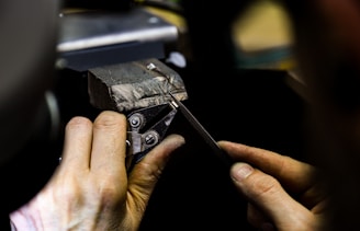 A person is using precision tools on a piece of metal secured in a vise, likely working on detailed craftsmanship or metalwork.