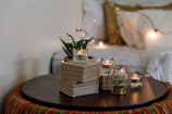 A cozy setting featuring a table with stacks of folded white napkins, a metal candle holder with a small plant, several tealight candles in glass and metal holders, and some string lights. The background shows a bed with fluffy pillows and a golden sequin cushion, enhancing the warm ambiance.