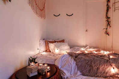 A cozy bedroom setup with a small bed covered in white and brown pillows and a fluffy, beige throw blanket. Soft string lights are draped across the bed, casting a warm glow. A round table beside the bed holds several lit candles, stacked books, and a small plant. Wall decor includes a hanging with tassels and fake eyelashes, alongside a door adorned with greenery and red flowers.