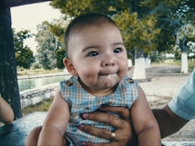 A baby with a plaid outfit sits outdoors, displaying a playful expression with cheeks puffed and lips pursed. An adult's hand supports the baby from the front. Trees and a stone bench by a water body form the serene backdrop.