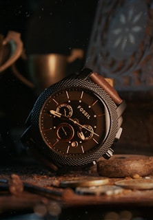 A sophisticated wristwatch with a textured metal bezel and brown leather strap is prominently displayed, surrounded by coins and a warm, dimly lit setting. The watch face displays multiple dials and the word 'FOSSIL' is visible.