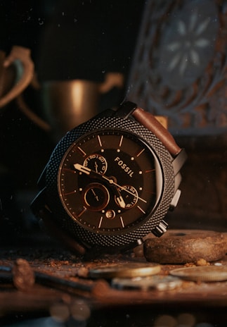 A sophisticated wristwatch with a textured metal bezel and brown leather strap is prominently displayed, surrounded by coins and a warm, dimly lit setting. The watch face displays multiple dials and the word 'FOSSIL' is visible.