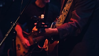 Two musicians are performing on stage, focused on guitar playing. One is wearing a jacket with a decorative strap featuring skull designs, while the other is playing an electric guitar beside a microphone stand. The setting is dimly lit, with a warm, intimate atmosphere.
