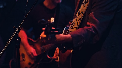 Two musicians are performing on stage, focused on guitar playing. One is wearing a jacket with a decorative strap featuring skull designs, while the other is playing an electric guitar beside a microphone stand. The setting is dimly lit, with a warm, intimate atmosphere.