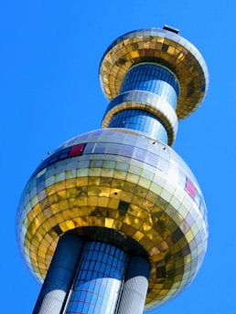 A modern, futuristic tower with a reflective surface, composed of large golden and silver spherical structures stacked vertically. The building's exterior is comprised of glass panels that mirror the bright blue sky.