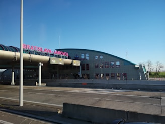 A modern building with the words 'BRATISLAVA JAROVCE' in red on the roof. The structure has a curved design and multiple windows, with a highway or road in the foreground and a clear blue sky. Trees line the horizon in the distance.