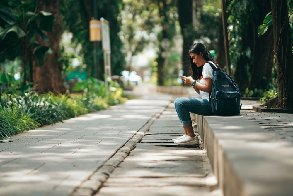 woman in black jacket and blue denim jeans sitting on concrete pathway during daytime