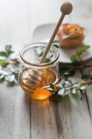 A glass jar filled with honey is placed on a wooden table. A wooden honey dipper rests inside the jar. Green branches are laid around the jar, and a pastry with apple slices is seen blurred in the background on a white plate.