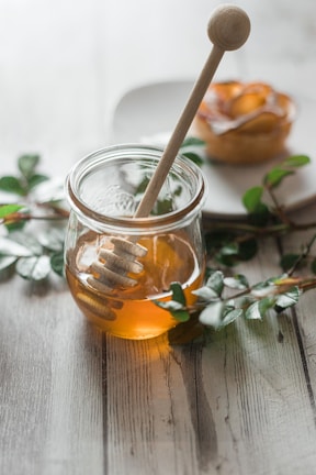 A glass jar filled with honey is placed on a wooden table. A wooden honey dipper rests inside the jar. Green branches are laid around the jar, and a pastry with apple slices is seen blurred in the background on a white plate.