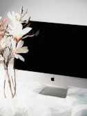 A sleek computer monitor with a blank black screen sits on a white desk. To the left, a clear vase holds several long-stemmed white and pink magnolia flowers.