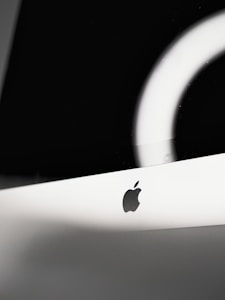 A sleek and minimalistic close-up of a computer monitor with the Apple logo prominently displayed on a white background. The lighting creates a modern and sophisticated ambiance with a reflective ring of light partially visible on the screen.