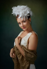 A woman wearing an elegant attire with a white feathered hat and holding a brown shawl. She has a subtle smile and elegant earrings, set against a softly lit, dark, and muted background.