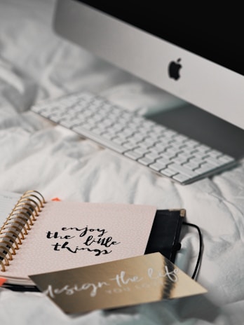 A desktop computer with a keyboard is positioned on a white, textured bedspread. In the foreground, a notebook with a spiral binding features the motivational phrase 'enjoy the little things' on its cover. Next to it lies a gold-colored card with the phrase 'design the life you love.'