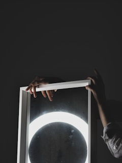 Hands holding a rectangular mirror with a bright circular light reflected on its surface, against a dark background.