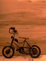A small, decorative metal bicycle with a wooden seat is positioned against a smooth, wooden background. The bicycle features two cylindrical handles and a classic design with visible pedal gears.