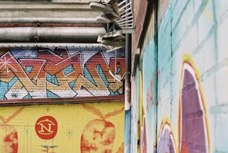 A series of graffiti-covered walls in an urban setting. The graffiti features a mix of abstract patterns and vibrant colors, predominantly reds, blues, and yellows. Multiple pairs of sneakers hang from the overhead pipes, adding an intriguing element to the scene.