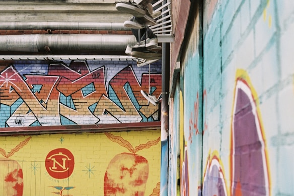 A series of graffiti-covered walls in an urban setting. The graffiti features a mix of abstract patterns and vibrant colors, predominantly reds, blues, and yellows. Multiple pairs of sneakers hang from the overhead pipes, adding an intriguing element to the scene.