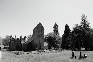A historic stone church surrounded by tall trees stands in the midst of a grassy field. Several people are walking and playing in the foreground. The sky is clear, enhancing the sharpness of the church's architectural features.