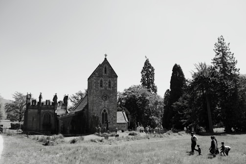A historic stone church surrounded by tall trees stands in the midst of a grassy field. Several people are walking and playing in the foreground. The sky is clear, enhancing the sharpness of the church's architectural features.