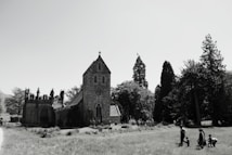 A historic stone church surrounded by tall trees stands in the midst of a grassy field. Several people are walking and playing in the foreground. The sky is clear, enhancing the sharpness of the church's architectural features.