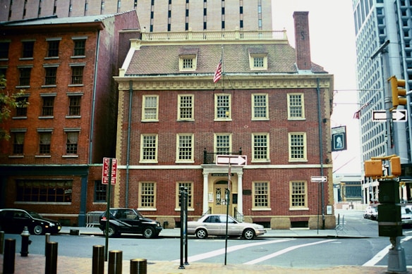 A historic red-brick building with multiple windows and an American flag mounted on the roof. The building is situated at a street intersection with a few cars parked along the curb. Street signs, traffic lights, and other city buildings surround the area, contributing to an urban setting.