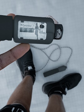 A hand holds a jump rope counter, displaying '0000' with a visible 'Counter' and 'Reset' button. Below, a person wears black athletic shoes with black socks. The jump rope lies on the ground in a loop.